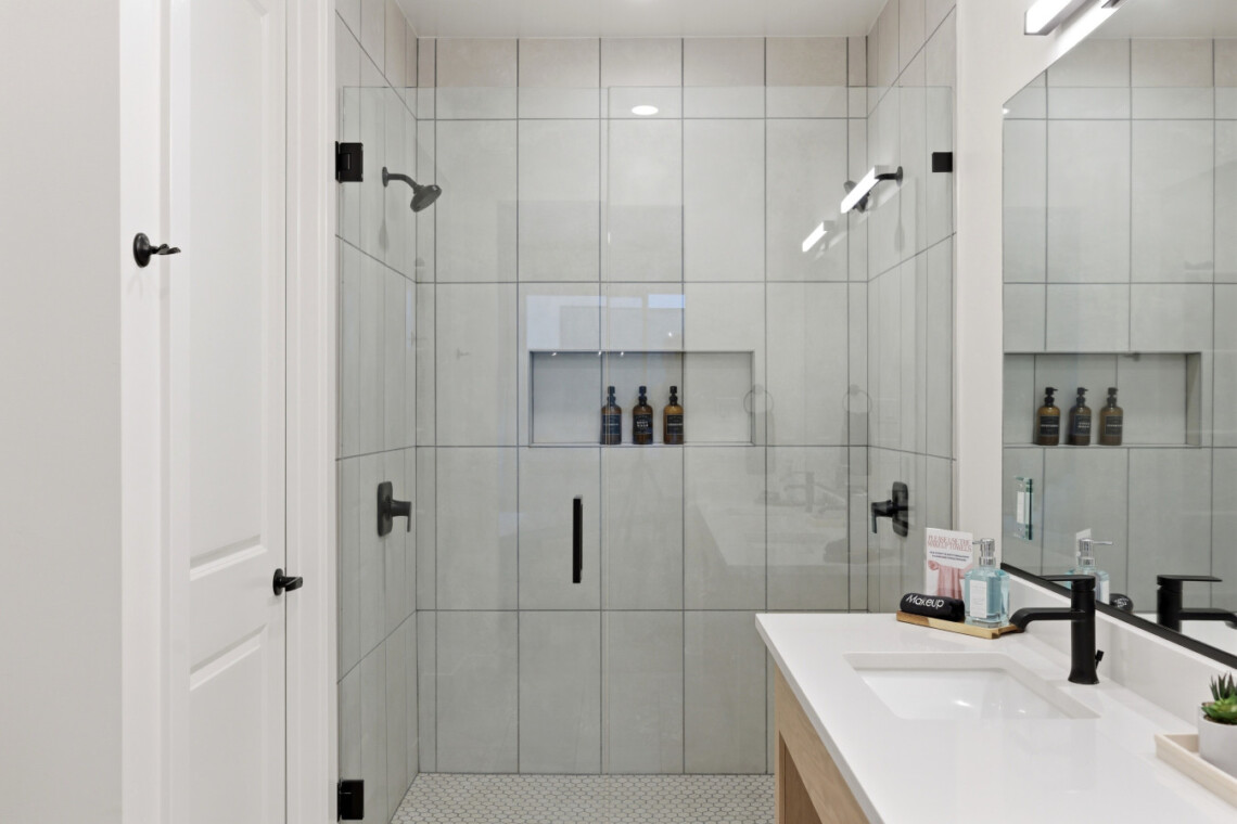 Modern glass-walled shower with built-in niches holding bottles, white tiled walls, and a white countertop sink with a black faucet.