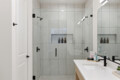 Modern glass-walled shower with built-in niches holding bottles, white tiled walls, and a white countertop sink with a black faucet.