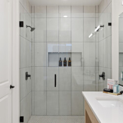Modern glass-walled shower with built-in niches holding bottles, white tiled walls, and a white countertop sink with a black faucet.