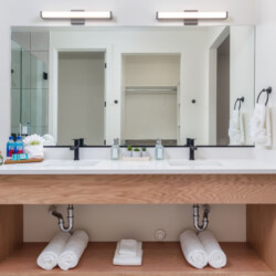 Double-sink bathroom vanity with white countertop, black faucets, and a large mirror above; open shelf underneath with rolled towels.
