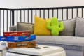 Gray sofa with a bright yellow pillow and a lime-green cactus plush, by a railing, with stacked board games (Yahtzee, Connect 4, Zion Opoly) on a white table in front.