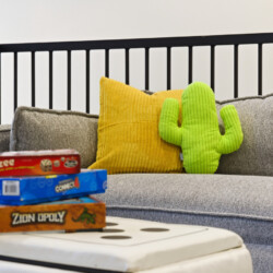 Gray sofa with a bright yellow pillow and a lime-green cactus plush, by a railing, with stacked board games (Yahtzee, Connect 4, Zion Opoly) on a white table in front.
