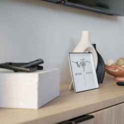 Framed white WiFi password card on a wooden console table, surrounded by vases, a decorative bowl, and remotes.