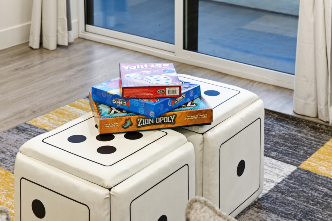Pile of family board games (Yahtzee, Connect 4, Zionopoly) on large white dice-pattern ottomans in a living room Chemin a window.