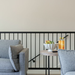 Grey living room with a round wooden table between a grey sofa and a grey armchair; a white bowl of snacks and two orange juice bottles sit on the table by a black metal railing.