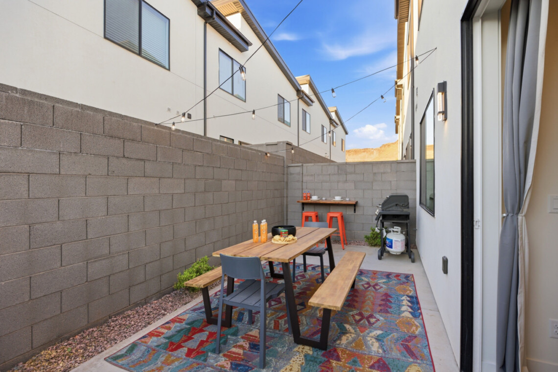 Backyard dining area with a wooden table and benches, colorful rug, string lights, and a grill against a gray block wall, outdoor sunset sky.