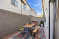 Backyard dining area with a wooden table and benches, colorful rug, string lights, and a grill against a gray block wall, outdoor sunset sky.