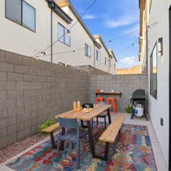 Backyard dining area with a wooden table and benches, colorful rug, string lights, and a grill against a gray block wall, outdoor sunset sky.