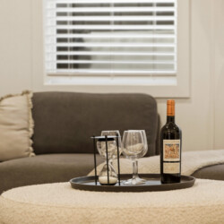 Wine bottle with two empty wine glasses and a small hourglass on a round black tray sitting on a soft, shaggy ottoman in a cozy living room.