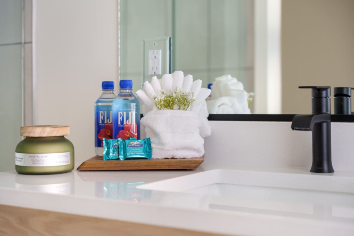Bathroom sink area with a wooden tray holding two Fiji water bottles, decorative blue soap wraps, white towels, and white flowers; modern black faucet in foreground.