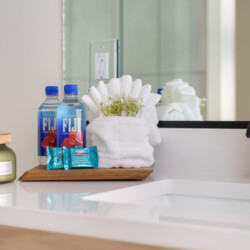 Bathroom sink area with a wooden tray holding two Fiji water bottles, decorative blue soap wraps, white towels, and white flowers; modern black faucet in foreground.