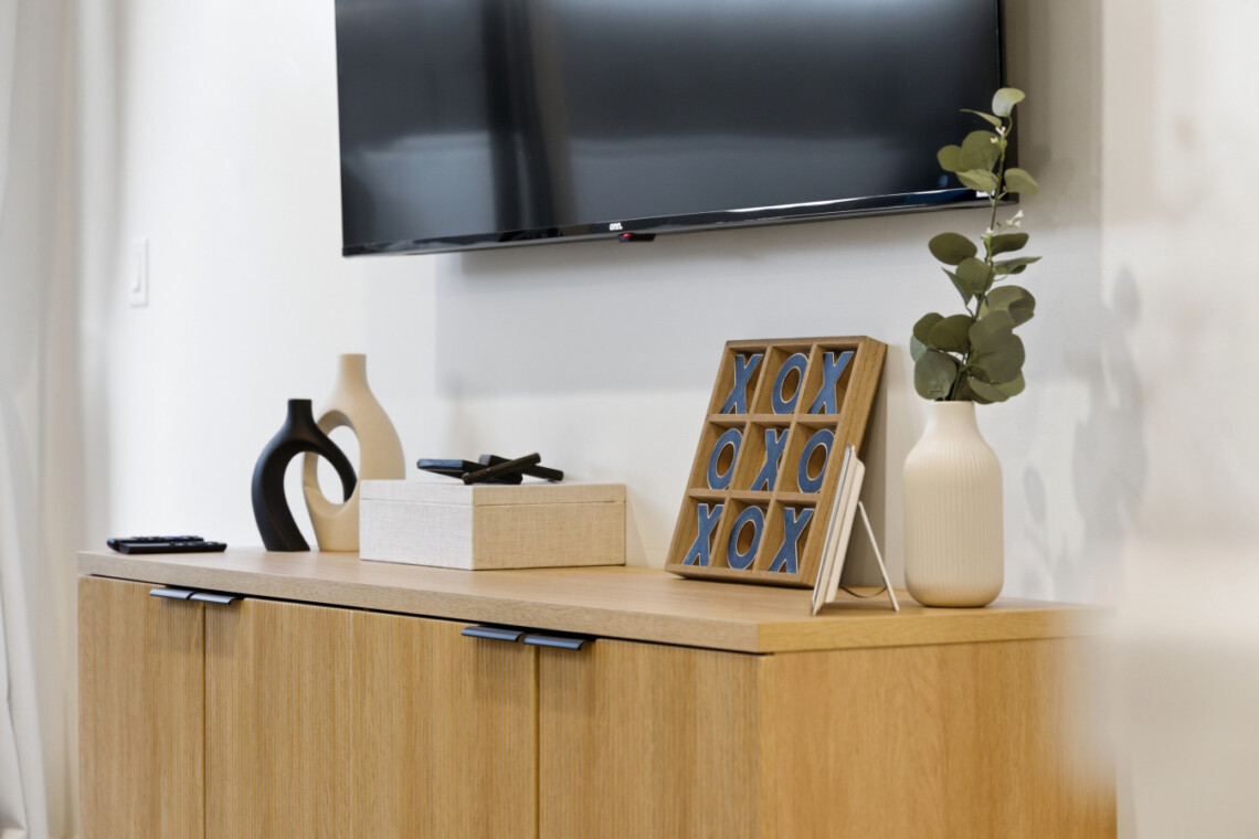 A wooden sideboard with decorative vases, a tic-tac-toe frame, a beige gift box, and a tall vase with eucalyptus under a wall-mounted TV.