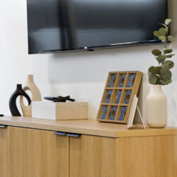 A wooden sideboard with decorative vases, a tic-tac-toe frame, a beige gift box, and a tall vase with eucalyptus under a wall-mounted TV.