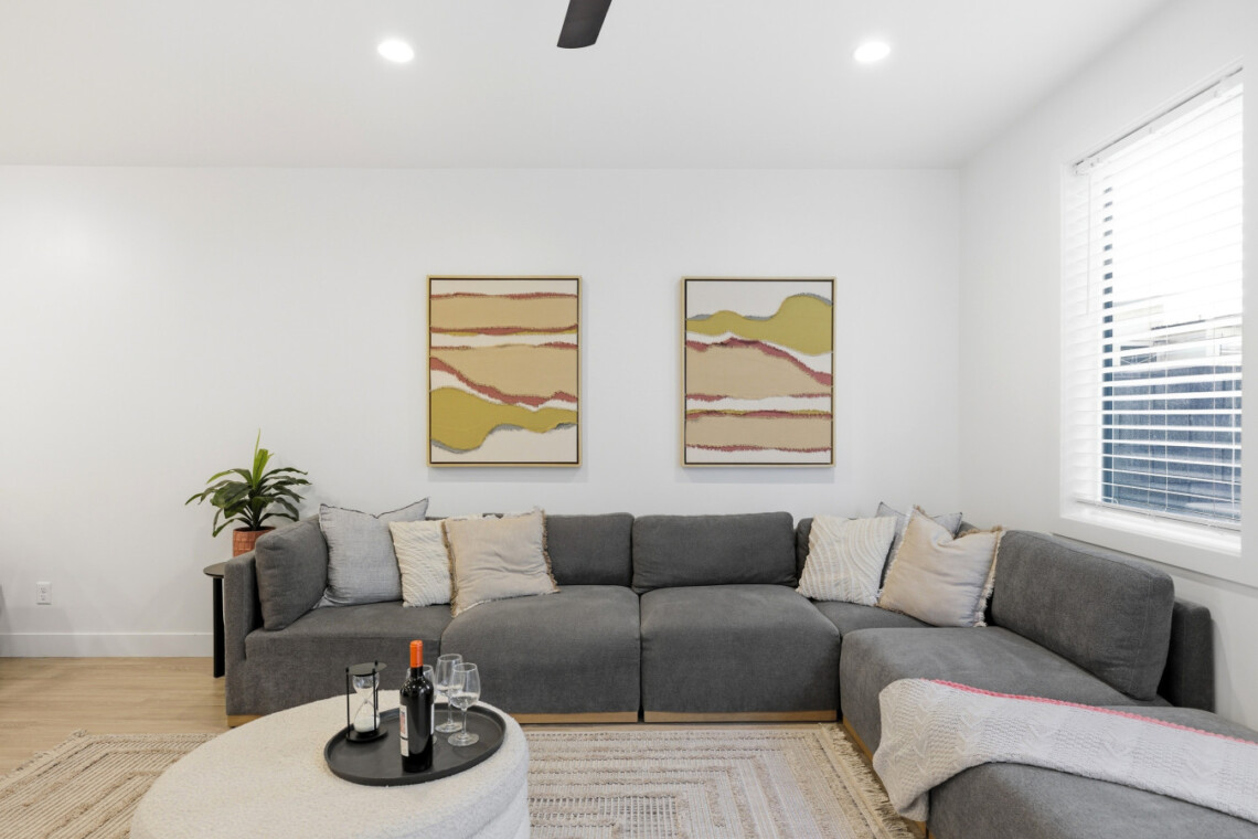 Grey L-shaped sofa with multiple neutral cushions in a bright living room, two abstract landscape paintings on the wall, and a round pouf-style coffee table with a wine bottle and glasses nearby.
