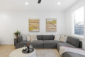 Grey L-shaped sofa with multiple neutral cushions in a bright living room, two abstract landscape paintings on the wall, and a round pouf-style coffee table with a wine bottle and glasses nearby.