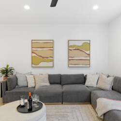 Grey L-shaped sofa with multiple neutral cushions in a bright living room, two abstract landscape paintings on the wall, and a round pouf-style coffee table with a wine bottle and glasses nearby.