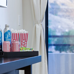 Tray of snacks on a dark table: two blue bottled drinks, two pink cans, a striped popcorn tub, and assorted candy wrappers near a window.