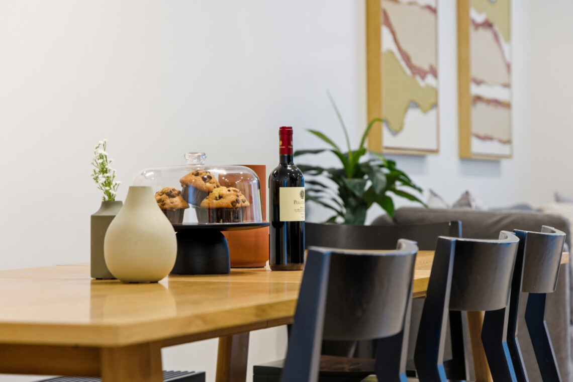 Wooden dining table with a glass cloche covering muffins, a bottle of red wine, and decorative vases/plant in a modern cafe setting.