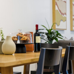 Wooden dining table with a glass cloche covering muffins, a bottle of red wine, and decorative vases/plant in a modern cafe setting.