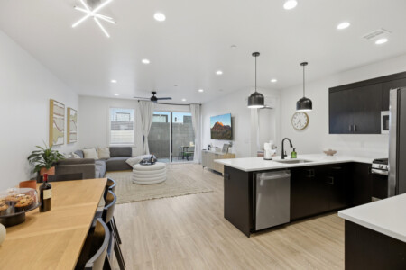 Open-concept living area with a gray sectional, round pouf and a coffee table, adjacent to a modern island kitchen with black cabinets and stainless appliances.