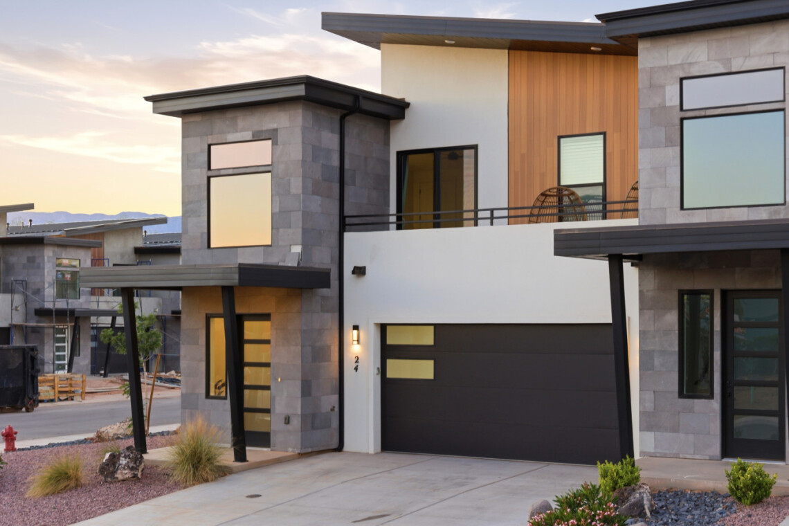 Modern two-story house exterior with gray stone panels, black garage door, wooden accent wall, and a balcony with hanging chairs at sunset.
