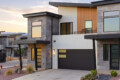 Modern two-story house exterior with gray stone panels, black garage door, wooden accent wall, and a balcony with hanging chairs at sunset.