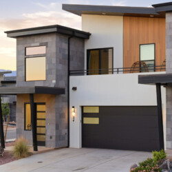 Modern two-story house exterior with gray stone panels, black garage door, wooden accent wall, and a balcony with hanging chairs at sunset.