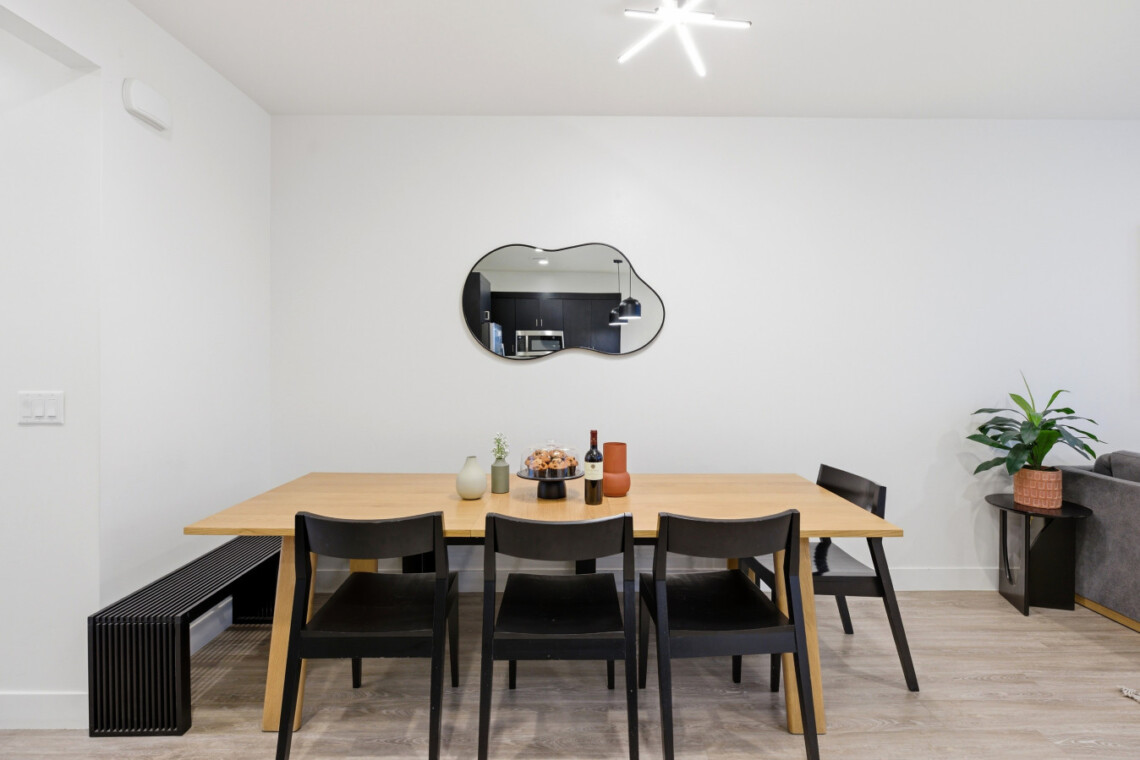 Modern dining room with a light wood table and six black chairs, a cloud-shaped mirror on a white wall, and a plant nearby.