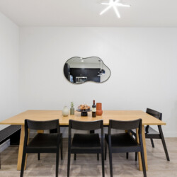 Modern dining room with a light wood table and six black chairs, a cloud-shaped mirror on a white wall, and a plant nearby.
