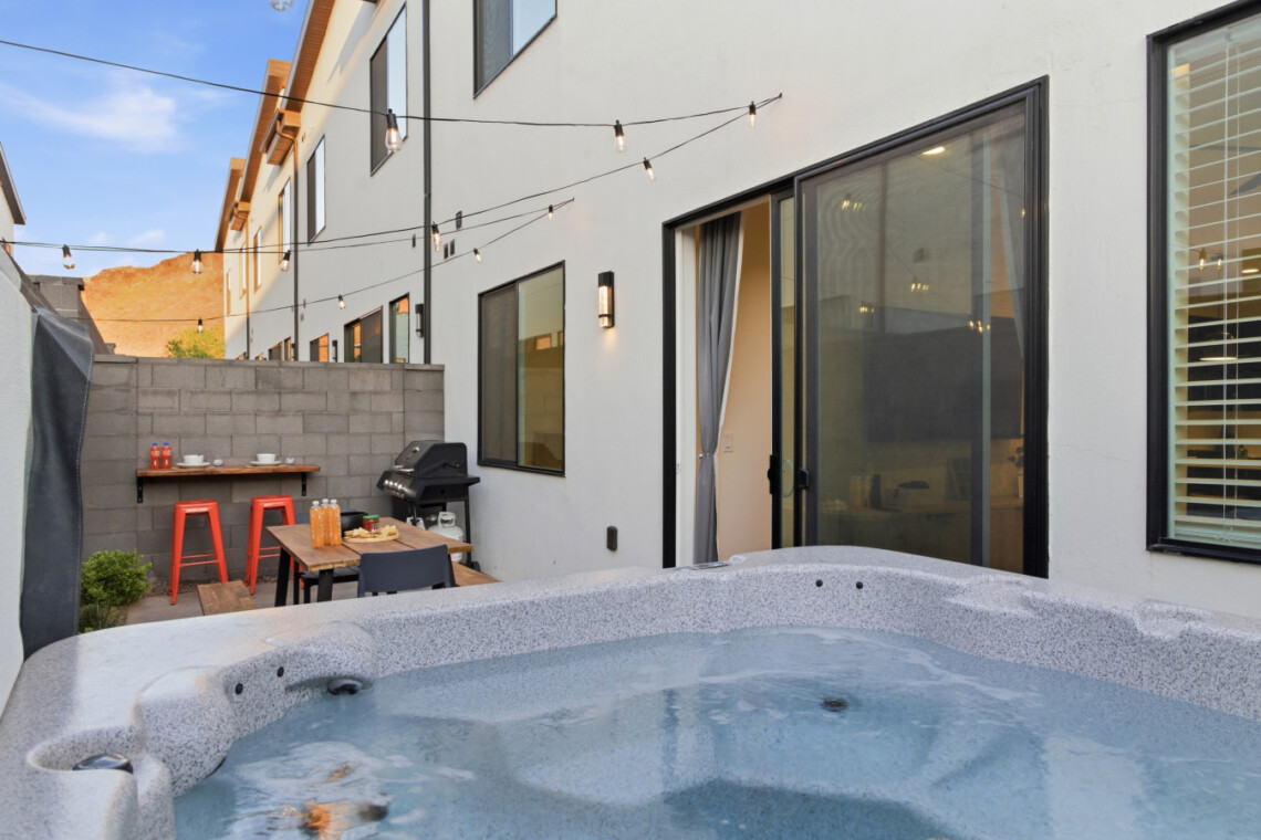 Backyard patio beside a white house with a hot tub in the foreground, string lights overhead, and a dining area with orange stools and a grill nearby.
