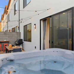 Backyard patio beside a white house with a hot tub in the foreground, string lights overhead, and a dining area with orange stools and a grill nearby.
