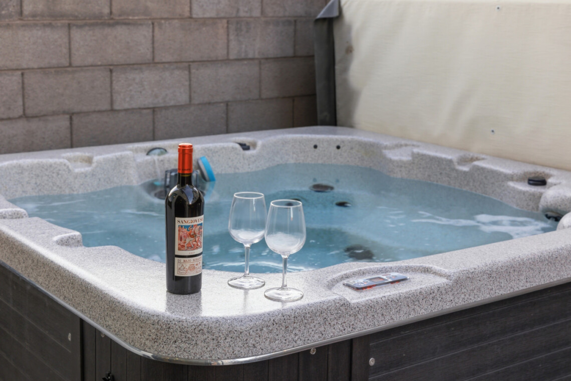 Wine bottle and two empty wine glasses on the edge of a hot tub in a backyard setting.