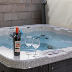 Wine bottle and two empty wine glasses on the edge of a hot tub in a backyard setting.