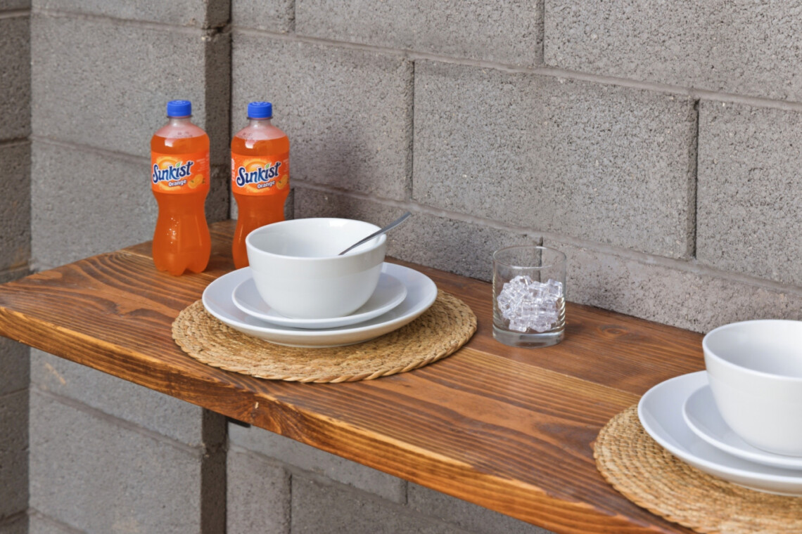 Outdoor wooden table set with white bowls and plates, two Sunkist orange soda bottles, a glass of ice, and a spoon on a brick wall backdrop.