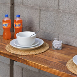Outdoor wooden table set with white bowls and plates, two Sunkist orange soda bottles, a glass of ice, and a spoon on a brick wall backdrop.