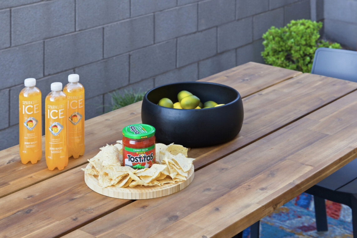 Outdoor wooden table with three Sparkling Ice bottles, a black bowl of limes, a jar of Tostitos salsa, and tortilla chips on a round board found in a backyard setup.