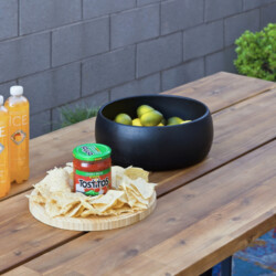 Outdoor wooden table with three Sparkling Ice bottles, a black bowl of limes, a jar of Tostitos salsa, and tortilla chips on a round board found in a backyard setup.