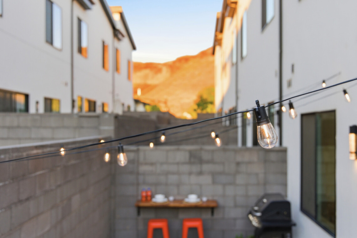 Cozy urban backyard with string lights, a wooden table set for three, and a barbecue grill between white modern houses at sunset.