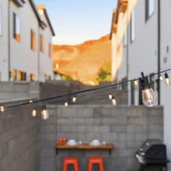 Cozy urban backyard with string lights, a wooden table set for three, and a barbecue grill between white modern houses at sunset.