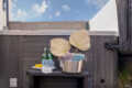 Relaxation scene on a wooden deck: a metal tub with rolled towels and large palm-leaf decor, next to two bottled waters and a lemon plate on a tray.