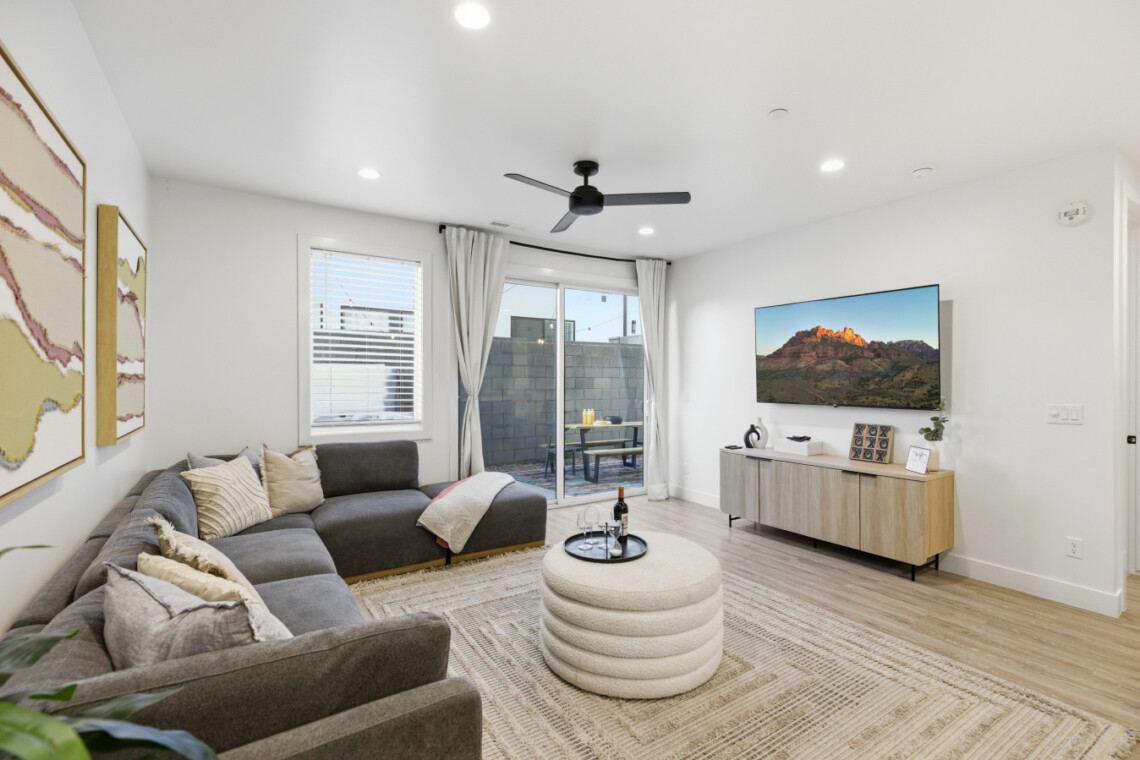 Modern living room with a gray sectional, neutral cushions, round beige ottoman, and a wooden-console TV setup above it.