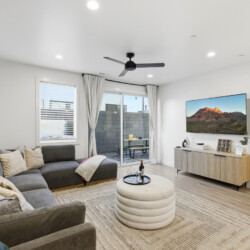 Modern living room with a gray sectional, neutral cushions, round beige ottoman, and a wooden-console TV setup above it.