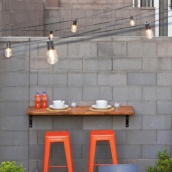 Outdoor patio scene with a wooden counter on a gray cinder block wall, two orange stools, place settings for two, string lights overhead, and a grill to the right.