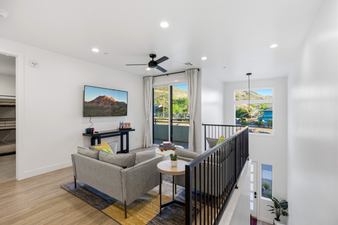 Modern living area with gray sofa, wall-mounted TV displaying a mountain scene, and a console table along a white wall; sliding glass door to a balcony beyond.