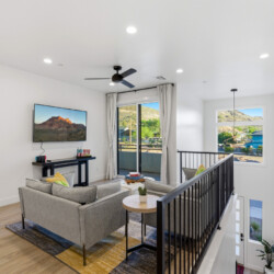 Modern living area with gray sofa, wall-mounted TV displaying a mountain scene, and a console table along a white wall; sliding glass door to a balcony beyond.