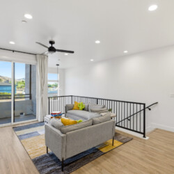Bright loft living room with gray sofa, colorful rug, and balcony door with white curtains; hillside view outside.