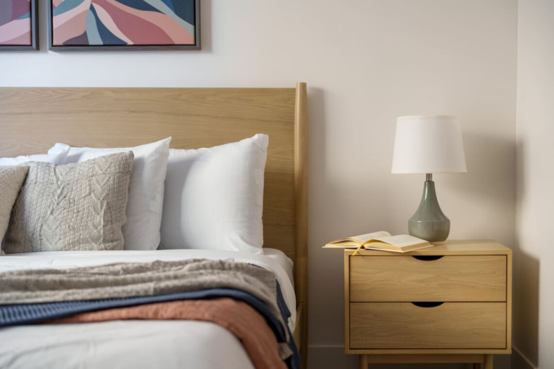 Bedroom scene with a wooden headboard, white pillows, and a folded blanket on the bed beside a beige nightstand with a lamp and an open book.