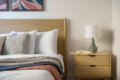 Bedroom scene with a wooden headboard, white pillows, and a folded blanket on the bed beside a beige nightstand with a lamp and an open book.