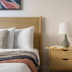 Bedroom scene with a wooden headboard, white pillows, and a folded blanket on the bed beside a beige nightstand with a lamp and an open book.