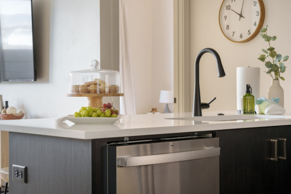 Contemporary kitchen island with a black gooseneck faucet, a glass cake stand with pastries, and a fruit platter on a white countertop.